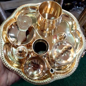 May include: A set of brass puja items, including a small plate, a cup, a spoon, a small pot, and a small incense burner. The items are arranged on a larger brass plate.