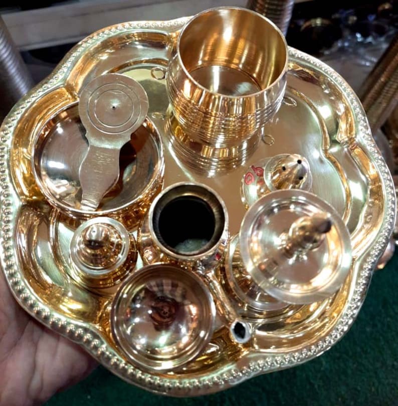 Op de afbeelding: Een set messing puja-artikelen, waaronder een klein bord, een beker, een lepel, een kleine pot en een kleine wierookbrander. De artikelen zijn gerangschikt op een groter messing bord.