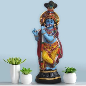 May include: A blue and gold statue of Krishna playing a flute. The statue is decorated with colourful jewels and beads. The statue is standing on a black base.