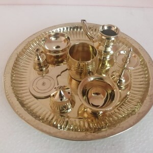 Op de afbeelding: Een set van acht gouden metalen puja-voorwerpen, waaronder een kleine kan, een kom, een bel en een bord, gerangschikt op een decoratieve dienblad.