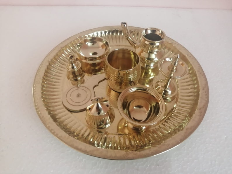 Op de afbeelding: Een set van acht gouden metalen puja-voorwerpen, waaronder een kleine kan, een kom, een bel en een bord, gerangschikt op een decoratieve dienblad.