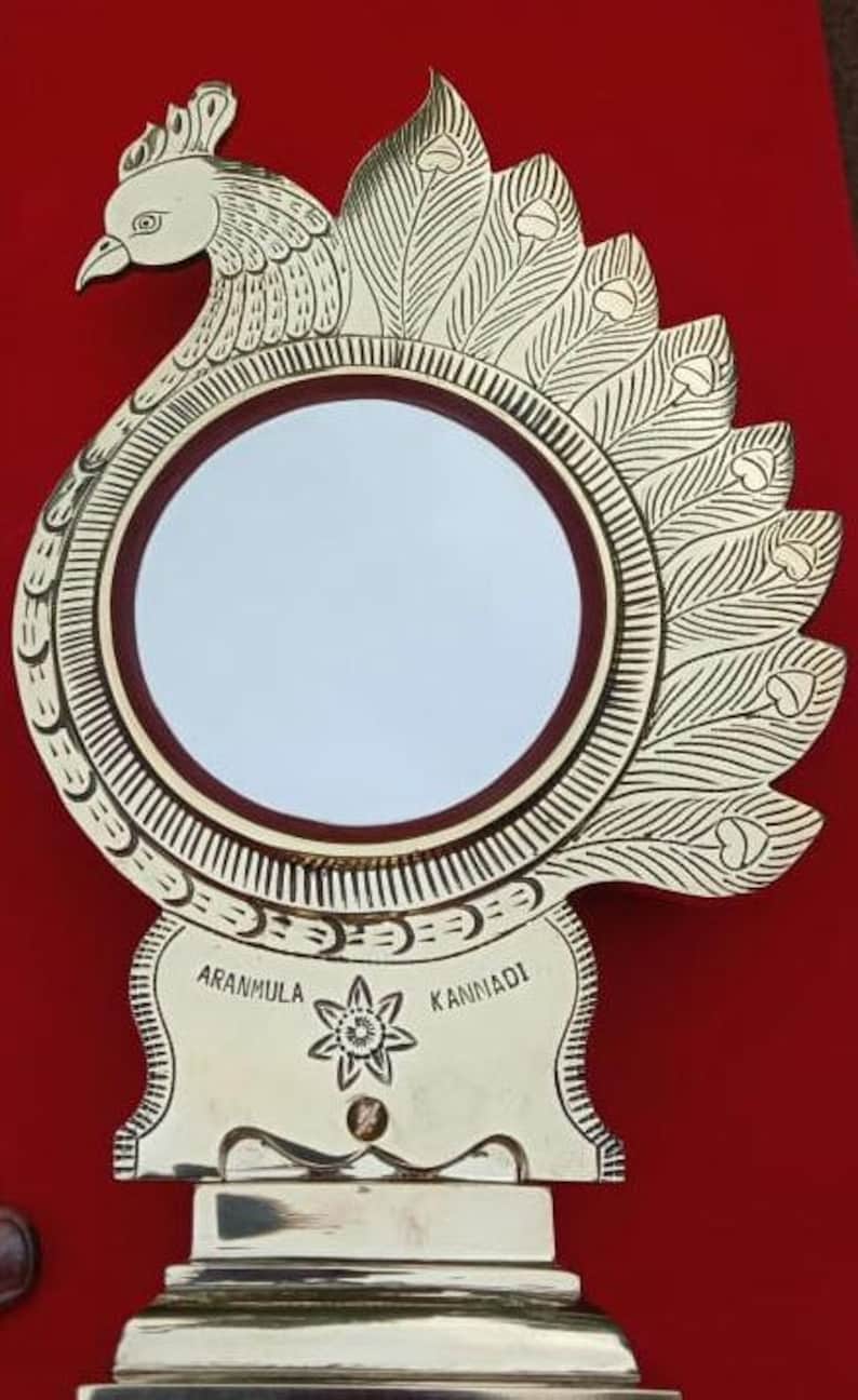 May include: A gold-colored peacock-shaped mirror with a round, reflective surface. The mirror is decorated with intricate details and the words "Aranmula Kannadi" are inscribed on the base.
