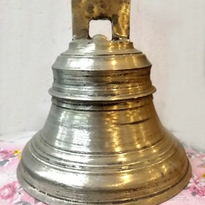 Puede incluir: Una campana de latón vintage con un asa rectangular simple. La campana tiene un acabado liso y envejecido y una forma ligeramente redondeada.