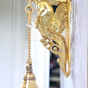 Peut inclure: Une sonnette de porte dorée ornée d'un motif de paon. La cloche est suspendue à un support décoratif avec des détails complexes. La cloche est attachée à une chaîne et est prête à être sonnée.