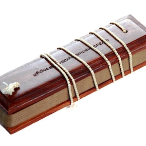 Puede incluir: Una caja de madera con un acabado marrón y una cuerda blanca atada alrededor. La caja tiene texto tamil en la parte superior que dice "சங்கீத சாஸ்திரம்".