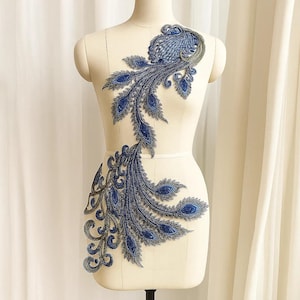 May include: A blue and gray embroidered peacock feather applique displayed on a mannequin. The applique features intricate detailing and a gradient of blue hues, with a touch of gray. The design is symmetrical and elegant, suitable for embellishing garments or crafts.