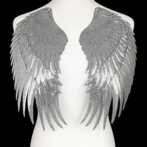 Puede incluir: Un par de alas de ángel plateadas con lentejuelas, exhibidas en un maniquí blanco. Las alas están detalladas con plumas en capas y lentejuelas brillantes, creando un efecto glamuroso y etéreo. Las alas son un artículo decorativo.