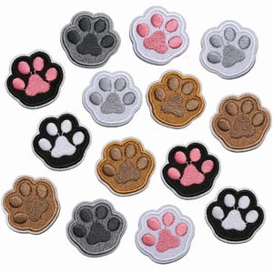 Op de afbeelding: Een verzameling geborduurde pootafdrukpatches in verschillende kleuren, waaronder roze, grijs, bruin en zwart. Elke patch heeft een gedetailleerd pootontwerp met een contrasterende rand, geschikt voor stoffen versiering.