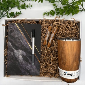 May include: A gift box filled with brown paper shred, a black and gold marble notebook, a silver pen, two brown candy sticks, and a brown wood-grain tumbler with the text "Swell" and "Tumbler" printed on the side.