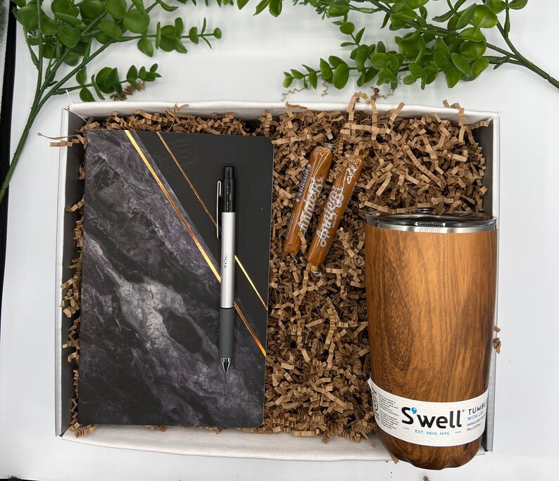 May include: A gift box filled with brown paper shred, a black and gold marble notebook, a silver pen, two brown candy sticks, and a brown wood-grain tumbler with the text "Swell" and "Tumbler" printed on the side.