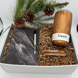 May include: A white gift box filled with brown shredded paper. The box contains a black and white marble notebook, a silver pen, two brown candy bars with white text, and a brown insulated tumbler with the text "Swell" in white.