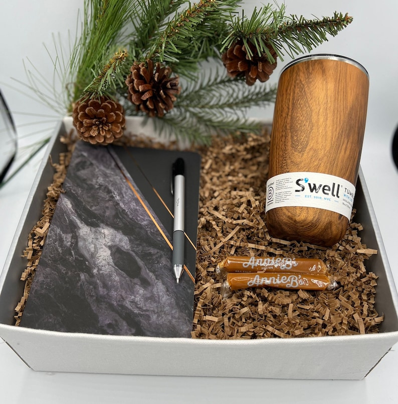 May include: A white gift box filled with brown shredded paper. The box contains a black and white marble notebook, a silver pen, two brown candy bars with white text, and a brown insulated tumbler with the text "Swell" in white.