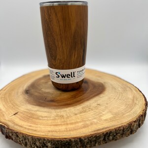 May include: A brown wood-grain stainless steel tumbler with a silver lid. The tumbler is sitting on a wooden slice. The tumbler has the text "Swell" and "Tumbler with Lid" on the side.