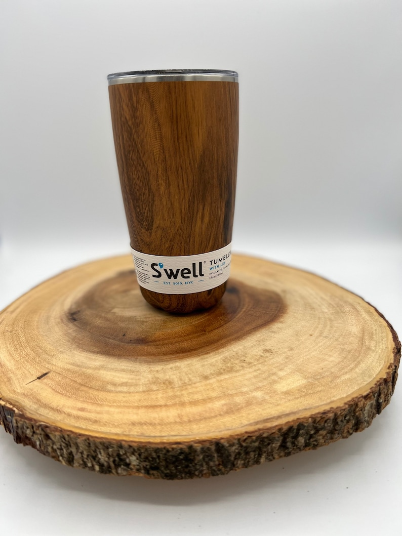 May include: A brown wood-grain stainless steel tumbler with a silver lid. The tumbler is sitting on a wooden slice. The tumbler has the text "Swell" and "Tumbler with Lid" on the side.