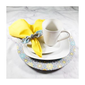 May include: A white ceramic mug with a handle sits on a white saucer. The saucer is on top of a larger, round, gray and white charger plate with a geometric pattern. A yellow napkin with a blue and green patterned trim is tied around the mug.