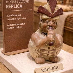 May include: A hand-stacked ceramic figurine replica from the Moche culture. The anthropomorphic vessel replica is beige and brown, with a detailed face and headdress. The figurine is holding a dark brown object to its mouth. The word "REPLICA" is on a small sign.
