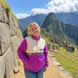 May include: A zip-up fleece jacket featuring a vibrant purple body and a cream-colored upper chest panel. The jacket includes a stand-up collar and horizontal bands decorated with a colorful geometric pattern across the chest and pockets.