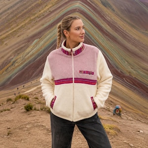 May include: A zip-up fleece jacket featuring a color-blocked design with a dusty rose upper section and cream lower section. The jacket includes a high collar, two side pockets, and a chest pocket, all accented with a colorful, patterned woven trim.