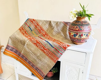 Op de afbeelding: Een beige en bruin geweven tapijt met kleurrijke strepen en geometrische patronen, gedrapeerd over een witte commode met een vaas met witte bloemen en een groene plant in een pot.