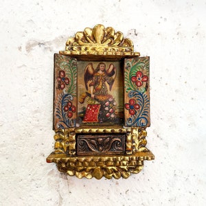 Puede incluir: Un pequeño santuario de madera con un acabado dorado y detalles florales. El santuario tiene una puerta con bisagras que se abre para revelar una imagen pintada de un ángel.