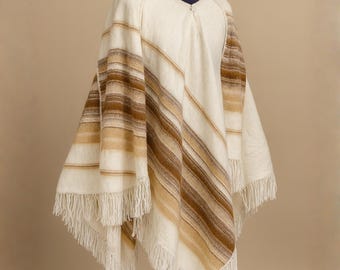 May include: Cream-colored poncho with brown and tan diagonal stripes. The poncho has a V-neck and fringe along the bottom edge. The garment is displayed on a mannequin with a navy blue turtleneck.