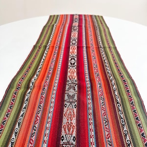 Puede incluir: Un colorido camino de mesa tejido con un diseño vibrante. El camino de mesa presenta rayas verticales en tonos de rojo, naranja, verde y blanco, con intrincados patrones geométricos. El camino de mesa está hecho de una tela gruesa y texturizada.