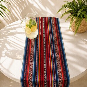 May include: A colorful table runner with a striped pattern in red, blue, and yellow. A glass pitcher filled with lemonade and mint sits on the runner. A potted fern is in the background.
