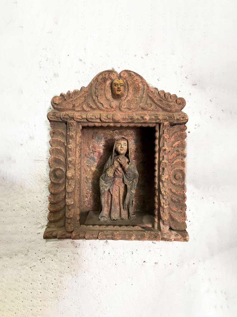 May include: A carved wooden frame with ornate details, featuring a small statue of a woman in a long robe holding a child. The frame is painted in shades of brown and red, with a yellow face on the top.