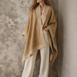 Puede incluir: Un poncho abierto de color beige con un borde ancho y texturizado de color crema en los extremos. La prenda se lleva sobre una camisa de botones de color claro y pantalones blancos holgados, combinados con zapatos de ante beige.