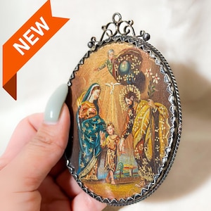 May include: Oval religious icon featuring a painted scene with figures in ornate robes, set within a decorative silver-toned frame. The artwork depicts a family with a child, with a "NEW" orange banner in the corner.