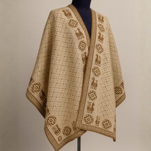 Puede incluir: Un poncho beige con un patrón geométrico y de llamas marrón. El poncho se exhibe en un maniquí y presenta un diseño de cuello en V. Los bordes están ribeteados con un borde marrón, y el estilo general recuerda a los textiles tradicionales sudamericanos.