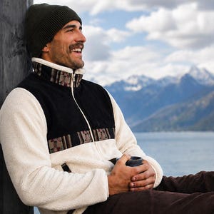 May include: A cream-coloured fleece jacket with a black upper chest panel and a patterned band featuring geometric shapes. The jacket has a high collar, a full-length front zip, and dark brown corduroy trousers. An olive green knit beanie is worn.