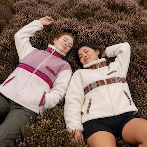 May include: Two zip-up fleece jackets with high collars and long sleeves. One jacket features a cream and mauve colour block design with a horizontal striped pattern. The other jacket is cream with a brown and cream patterned horizontal band.