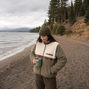 May include: A full-zip fleece jacket featuring olive green sleeves and back, a cream-colored chest panel, and a decorative patterned trim across the chest and pockets. The jacket is paired with a matching olive green beanie and a sage green tumbler.