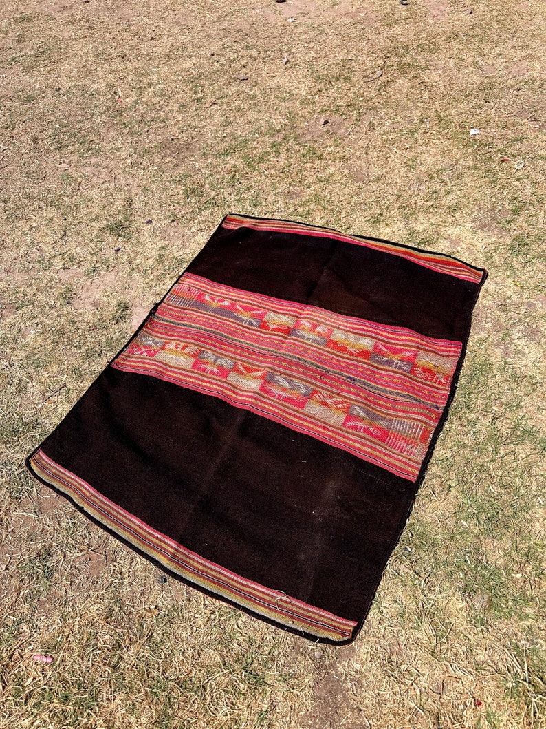 Op de afbeelding: Een zwart en rood geweven alpaca wollen deken met een geometrisch patroon. Het deken ligt op een bed van droog gras.
