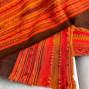 K&ouml;nnte beinhalten: Ein gewebtes Textil mit braunem Hintergrund und bunten Streifen in Orange-, Rot- und Gelbt&ouml;nen. Das Textil hat ein traditionelles Muster und ist aus einem weichen, strukturierten Material gefertigt.