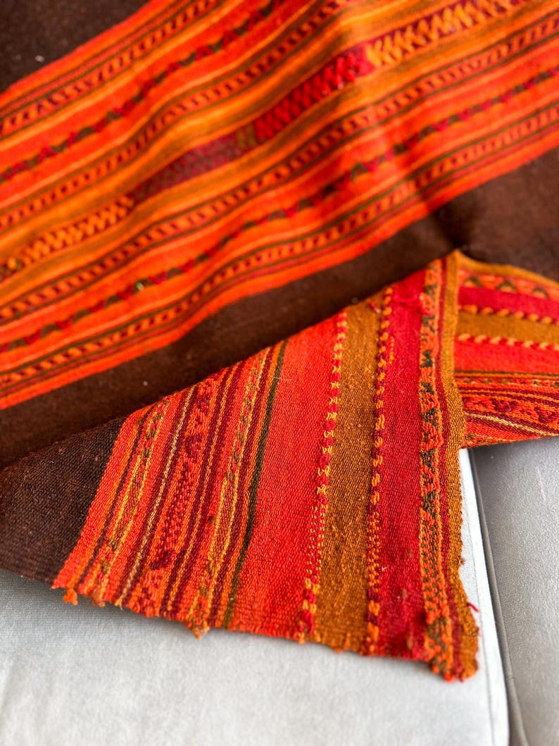K&ouml;nnte beinhalten: Ein gewebtes Textil mit braunem Hintergrund und bunten Streifen in Orange-, Rot- und Gelbt&ouml;nen. Das Textil hat ein traditionelles Muster und ist aus einem weichen, strukturierten Material gefertigt.