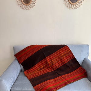 K&ouml;nnte beinhalten: Eine braune, rote und goldene gestreifte Wolldecke mit komplizierten Mustern liegt &uuml;ber einem hellgrauen Sofa. Die Decke ist aus einem weichen, gewebten Stoff und hat ein rustikales, bohemes Flair.