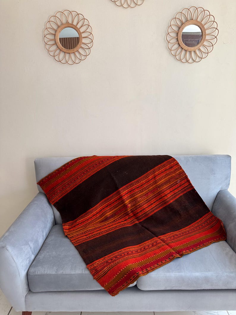 K&ouml;nnte beinhalten: Eine braune, rote und goldene gestreifte Wolldecke mit komplizierten Mustern liegt &uuml;ber einem hellgrauen Sofa. Die Decke ist aus einem weichen, gewebten Stoff und hat ein rustikales, bohemes Flair.