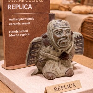 May include: A hand-crafted Moche culture replica ceramic vessel. The figure is a seated anthropomorphic form with detailed facial features and patterned wings. The sign reads "Moche Culture Replica".