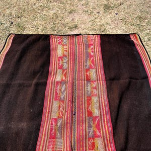 Op de afbeelding: Een zwarte alpaca wollen deken met een kleurrijke geweven rand. De rand heeft geometrische patronen in rood, geel en blauw.