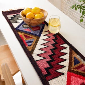 Puede incluir: Un camino de mesa tejido de colores con patrones geométricos en rojo, azul, marrón y blanco. Un cuenco de madera lleno de naranjas y un vaso de agua con rodajas de limón están sobre la mesa.