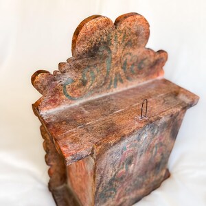 May include: A small wooden box with a carved, ornate top. The box is painted in shades of brown, red, and green, with a decorative floral design. The box has a small metal hook on the top.