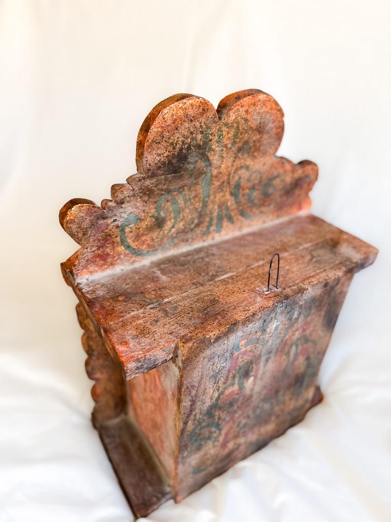 May include: A small wooden box with a carved, ornate top. The box is painted in shades of brown, red, and green, with a decorative floral design. The box has a small metal hook on the top.