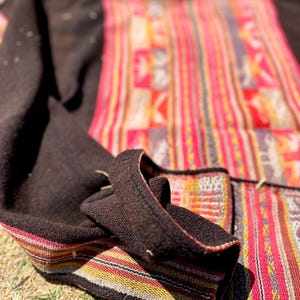 Op de afbeelding: Een kleurrijk geweven textiel met een zwarte rand. Het textiel heeft een gestreept patroon met rode, gele en witte strepen. Het textiel is dubbelgevouwen en ligt op een bed van droog gras.