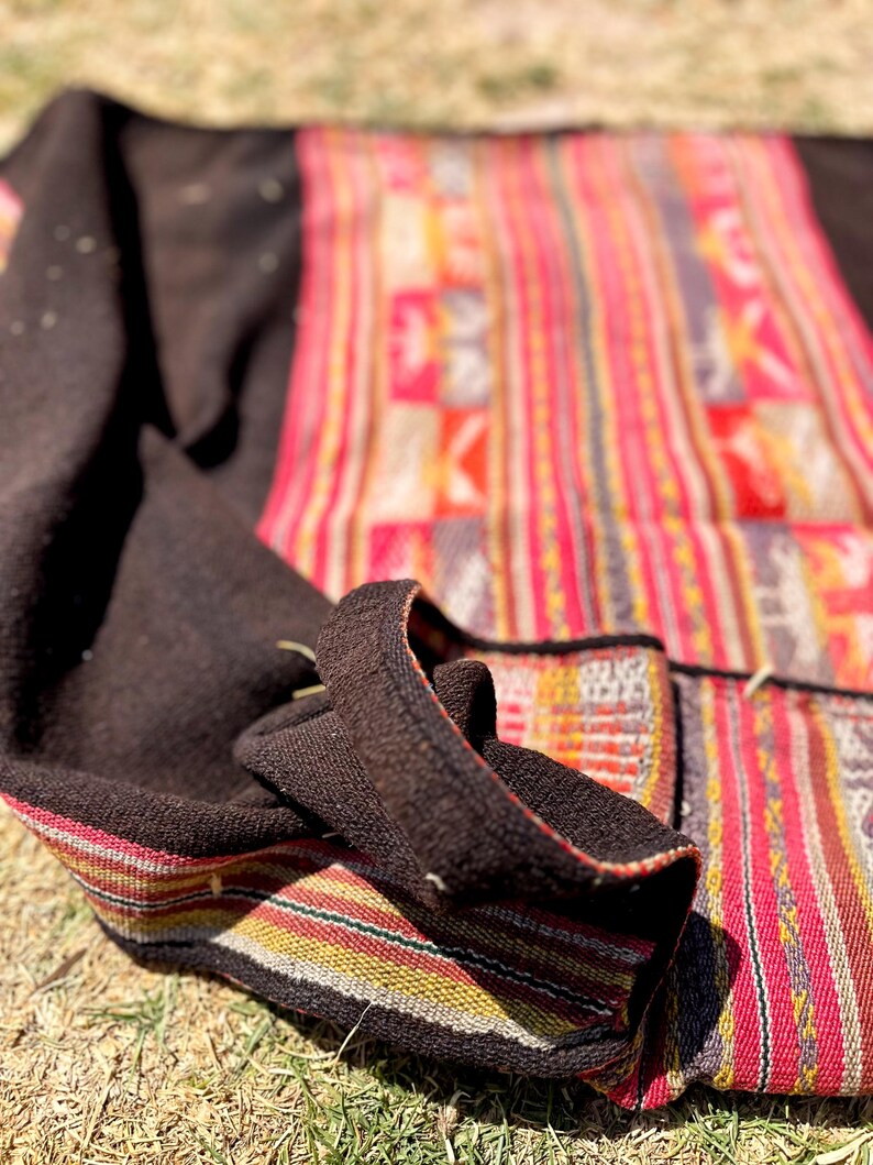 Op de afbeelding: Een kleurrijk geweven textiel met een zwarte rand. Het textiel heeft een gestreept patroon met rode, gele en witte strepen. Het textiel is dubbelgevouwen en ligt op een bed van droog gras.