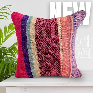 May include: A square throw cushion featuring a woven, multi-coloured striped pattern in shades of magenta, purple, teal, beige, and coral. The fabric has a textured, coarse weave. The text NEW ARRIVAL is visible in the background in white letters.