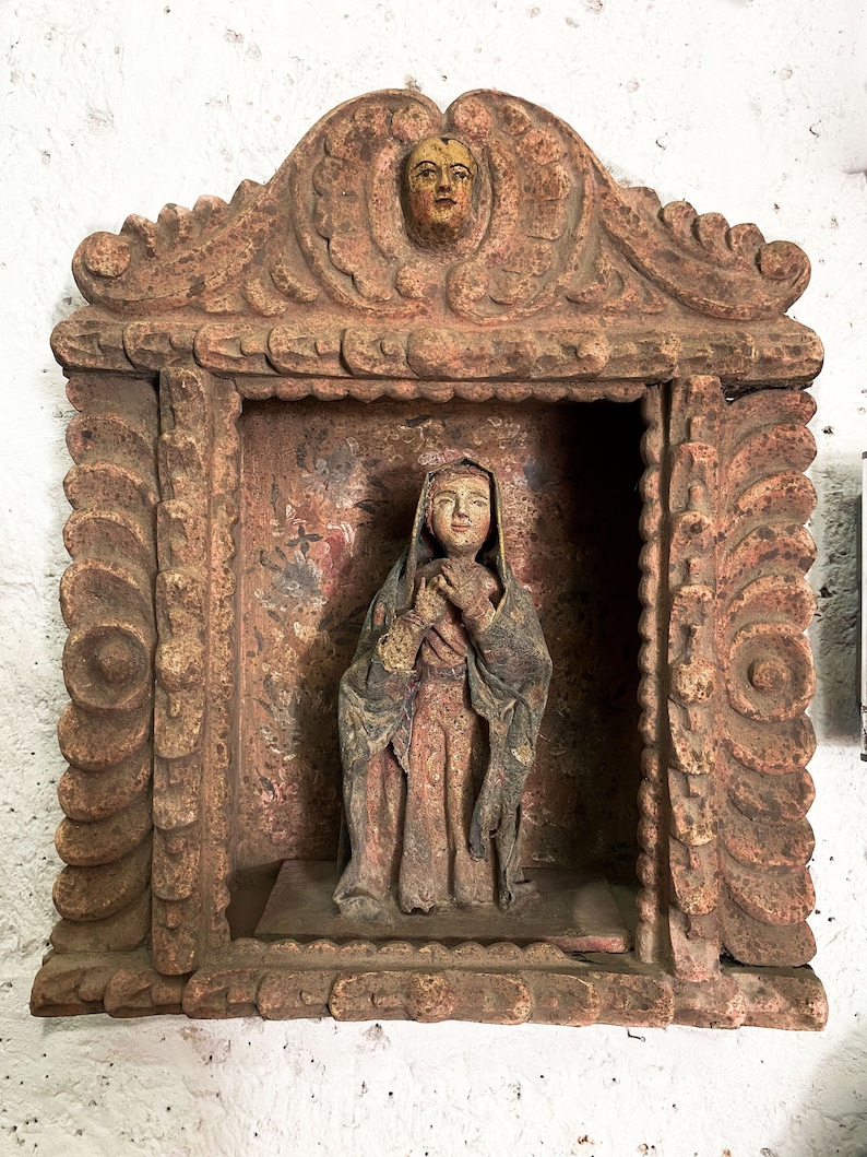 May include: A carved wooden frame with a detailed design, featuring a small statue of a woman in a long robe, with a painted face and hands. The frame is painted in shades of brown and red, with a small painted face at the top.