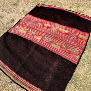 Op de afbeelding: Een zwart en bruin geweven deken met kleurrijke strepen en geometrische patronen. De deken is gemaakt van alpaca wol en is een traditioneel Peruaans textiel.