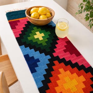 May include: A vibrant, patterned table runner in blue, green, pink, orange, and black. A wooden bowl of lemons and a glass of lemonade sit on a white table. The runner's geometric design adds a splash of colour to the setting, complementing the fresh fruit.
